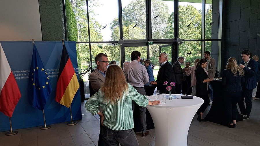Deutsche Botschaft, Round Table: Teilnehmerinnen und Teilnehmer in Gesprächen an verschiedenen Stehtischen. Im Hintergrund Fahnen EU, Deutschland und Polen.