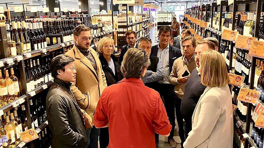 Ein Gruppe von Menschen steht in einem Supermarkt zwischen Weinregalen.